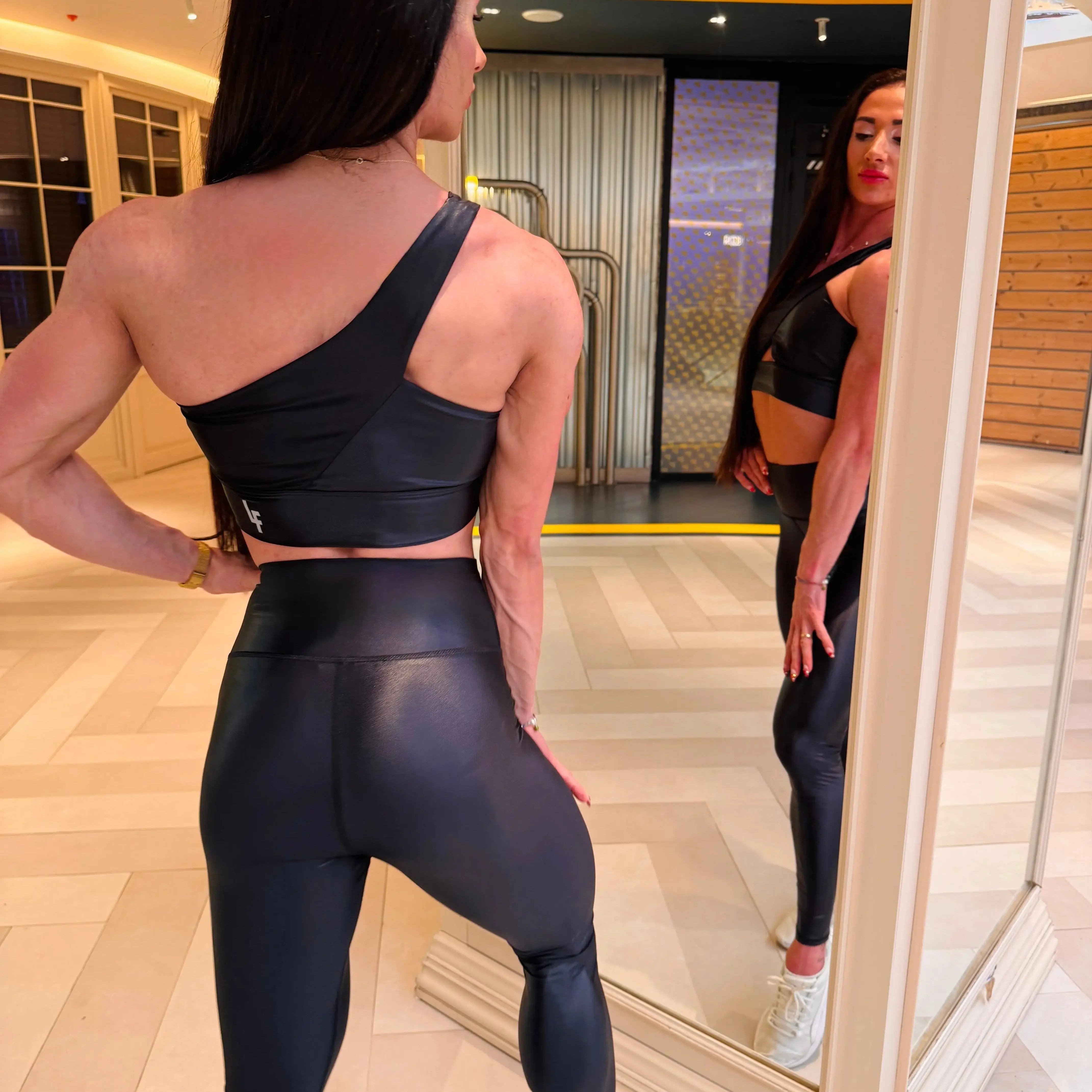 Mujer luciendo el top asimétrico brillante Top Carmela Pearl Shine de LUXFEEL en color Negro Brillante, con diseño de un solo hombro y apertura en el escote.