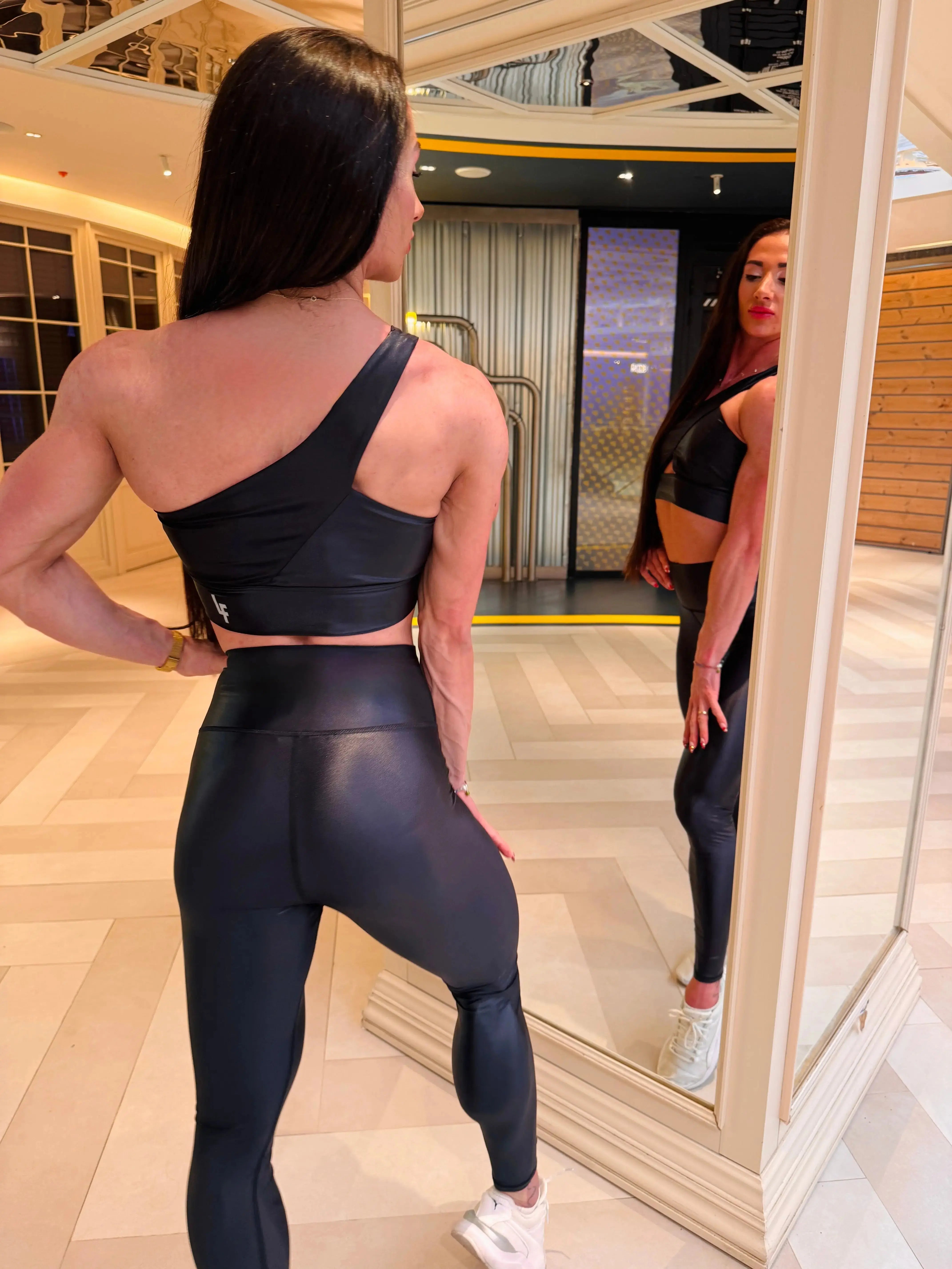 Mujer luciendo el top asimétrico brillante Top Carmela Pearl Shine de LUXFEEL en color Negro Brillante, con diseño de un solo hombro y apertura en el escote.