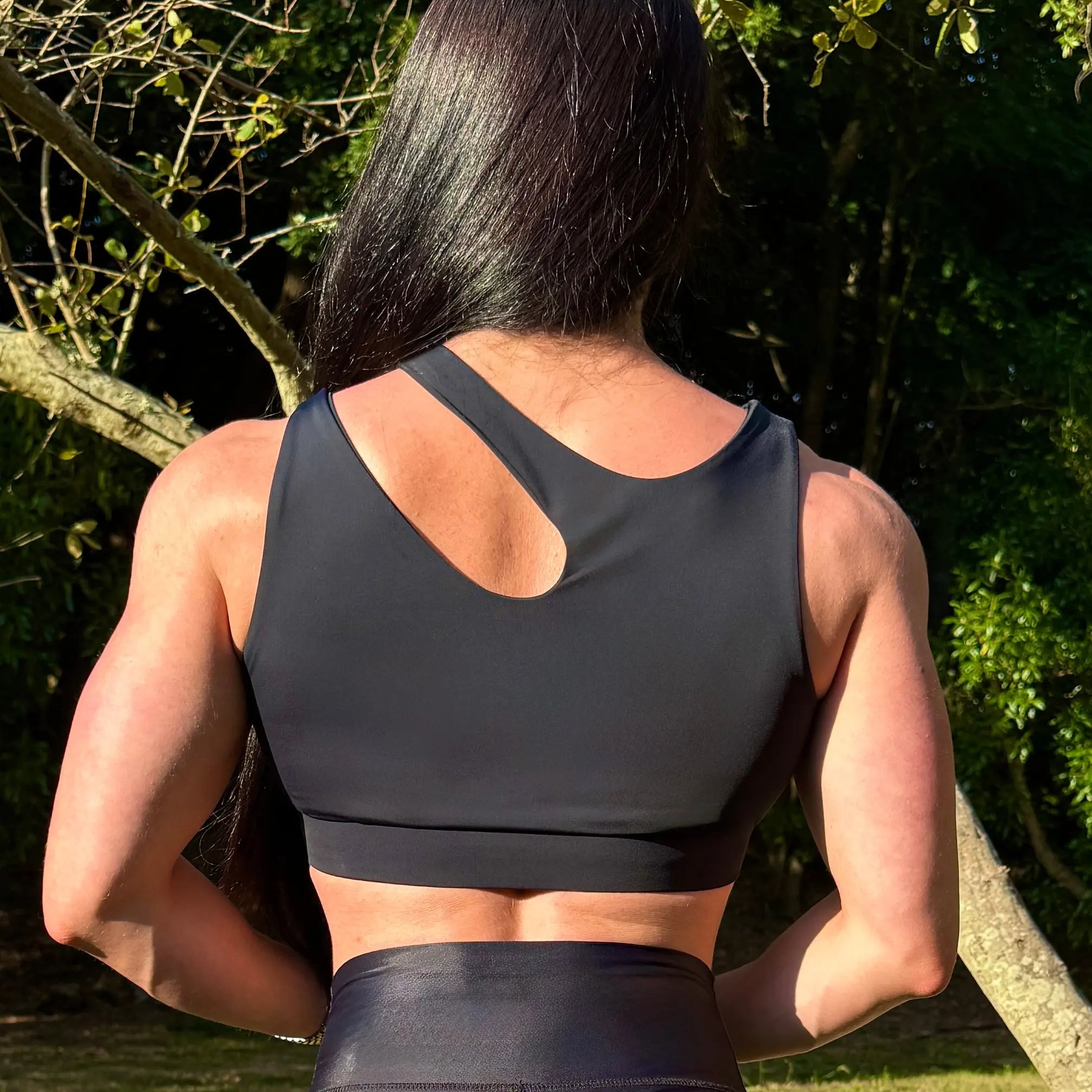 Modelo en la naturaleza mostrando Top Vanesa (parte trasera) de LUXFEEL en color negro, con un diseño de apertura en el hombro y soporte extra.