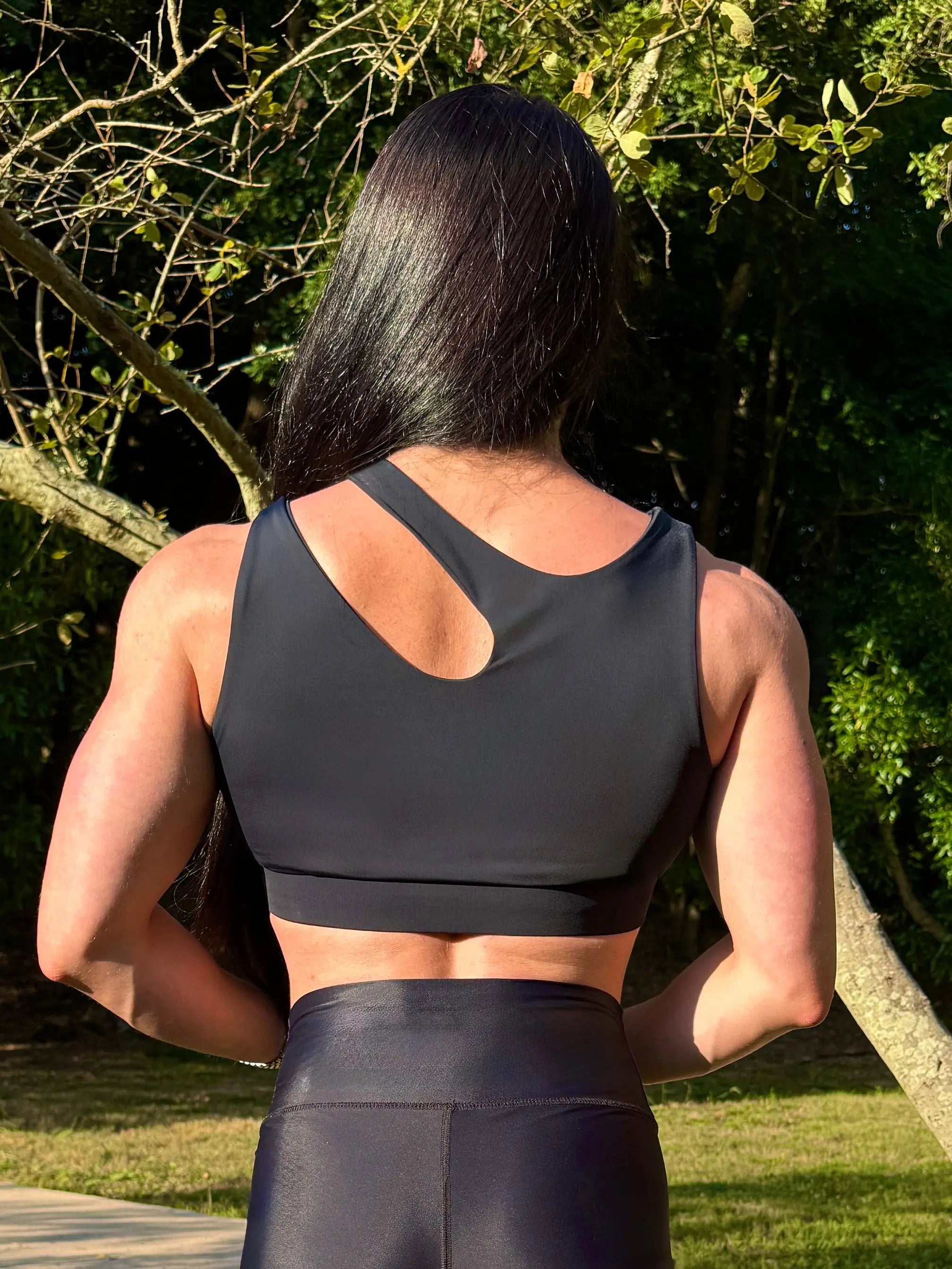 Modelo en la naturaleza mostrando Top Vanesa (parte trasera) de LUXFEEL en color negro,  con un diseño de apertura en el hombro y soporte extra.